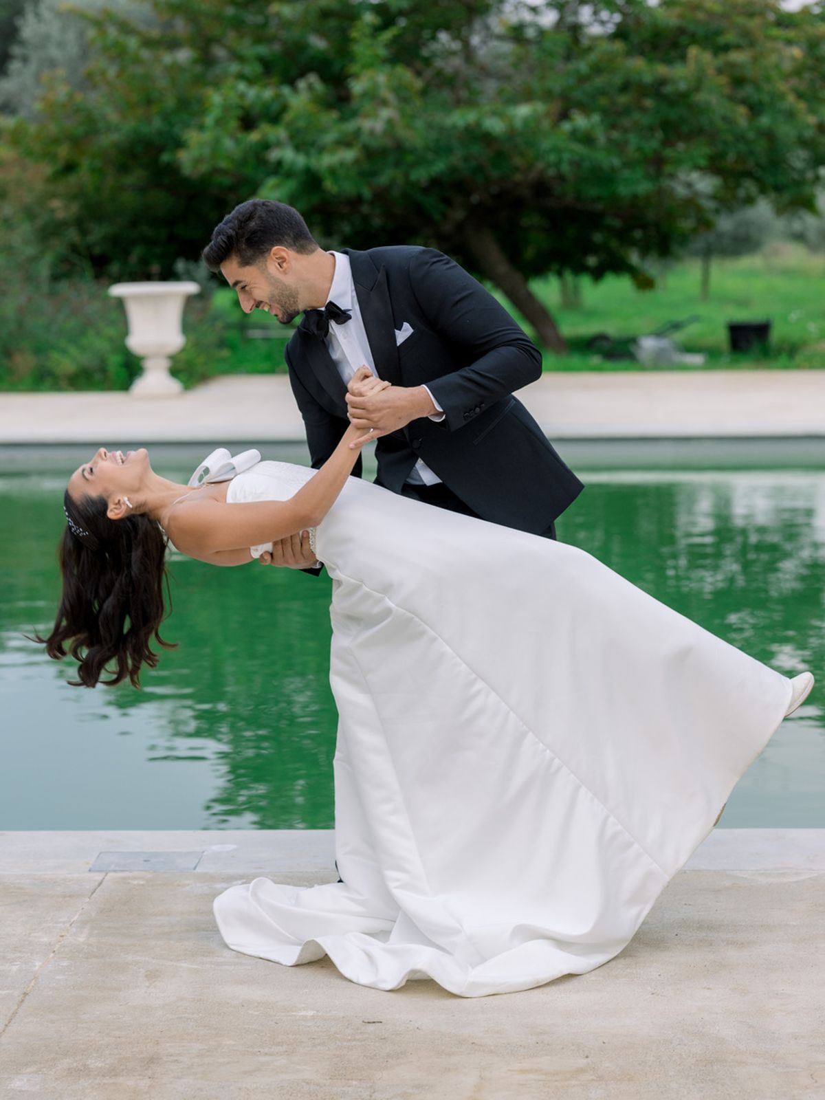 Groom dipping laughing bride beside a reflecting pool in a garden setting with ornamental planters in the background