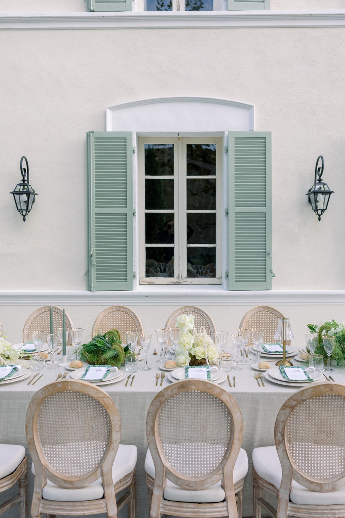 Long table with sage candles, ivory roses, and cane-back chairs against white facade with green shutters