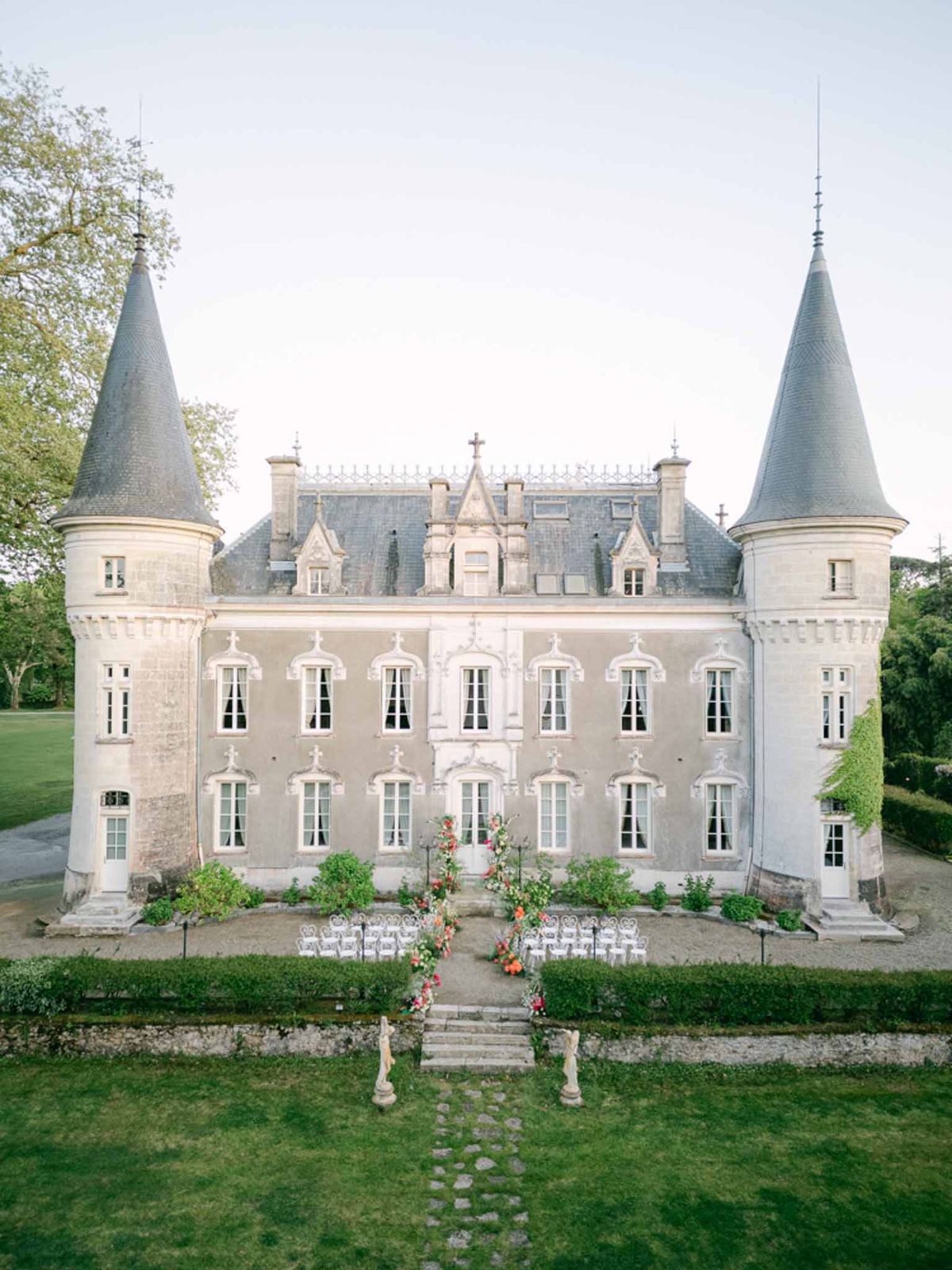 Aerial view of French chateau with slate turrets and outdoor ceremony setup with coral floral arch and white chairs