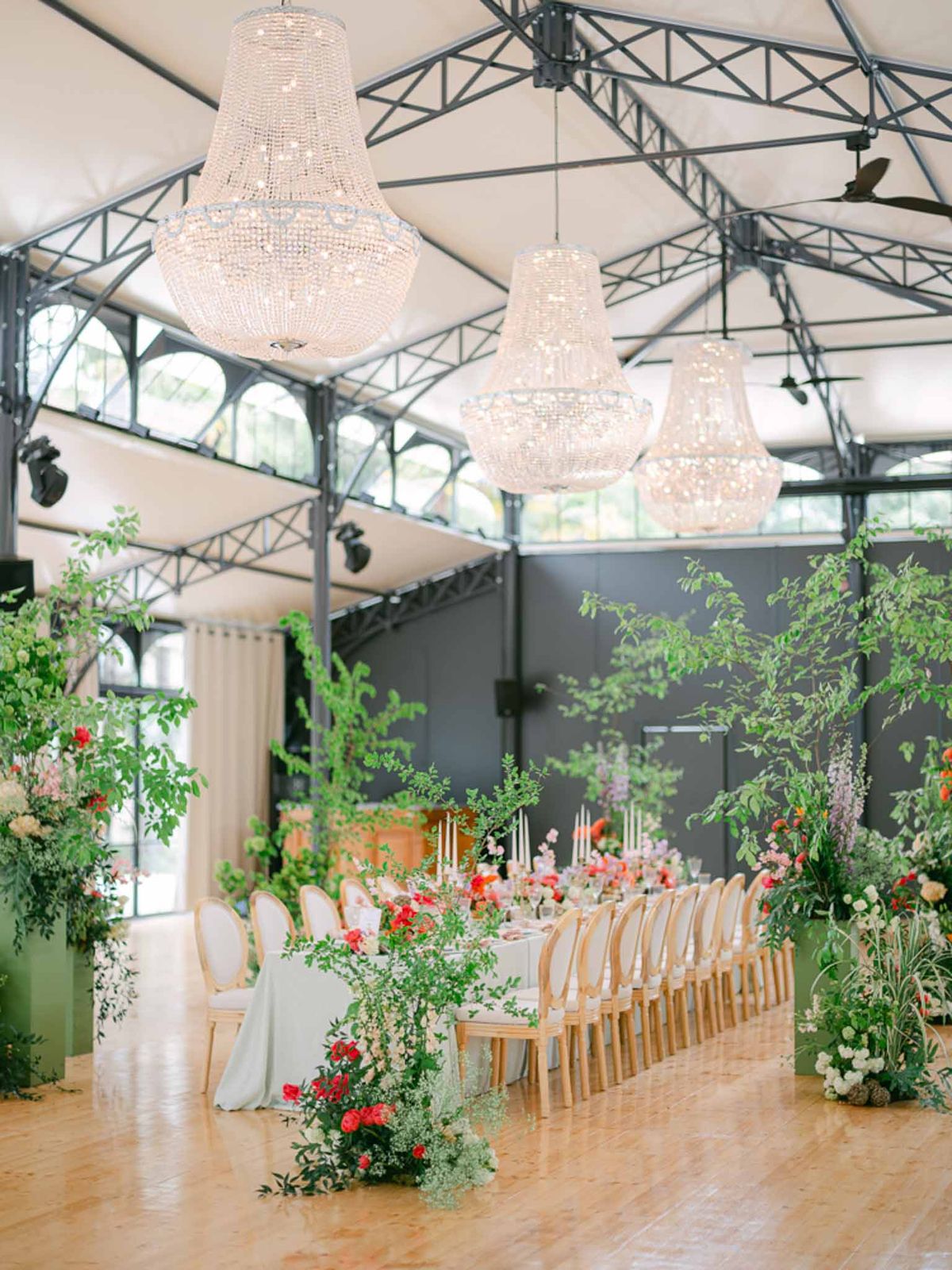 Greenhouse reception with crystal chandeliers, long table with red and coral floral centerpieces, and potted trees