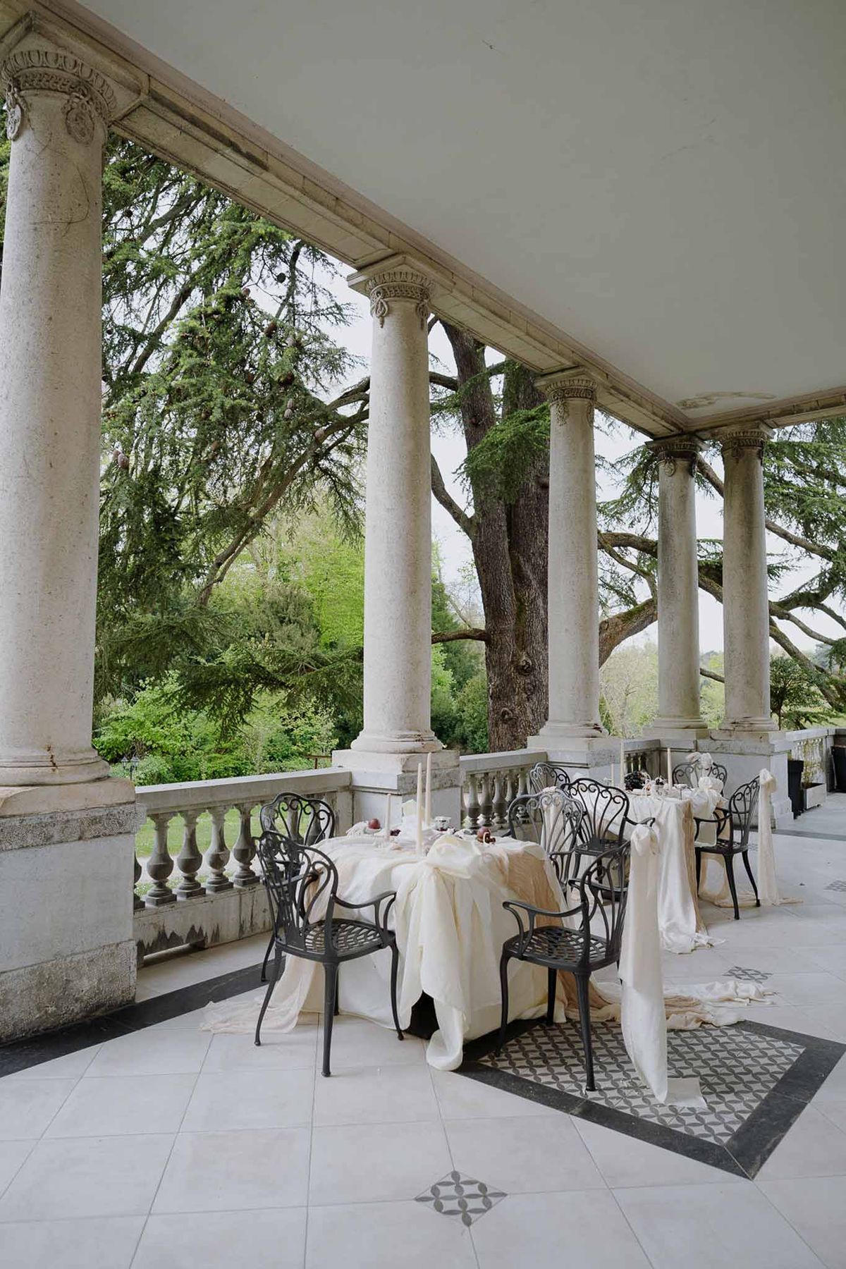 Colonnade terrace with Corinthian columns, wrought-iron chairs, and ivory draped tables overlooking parkland