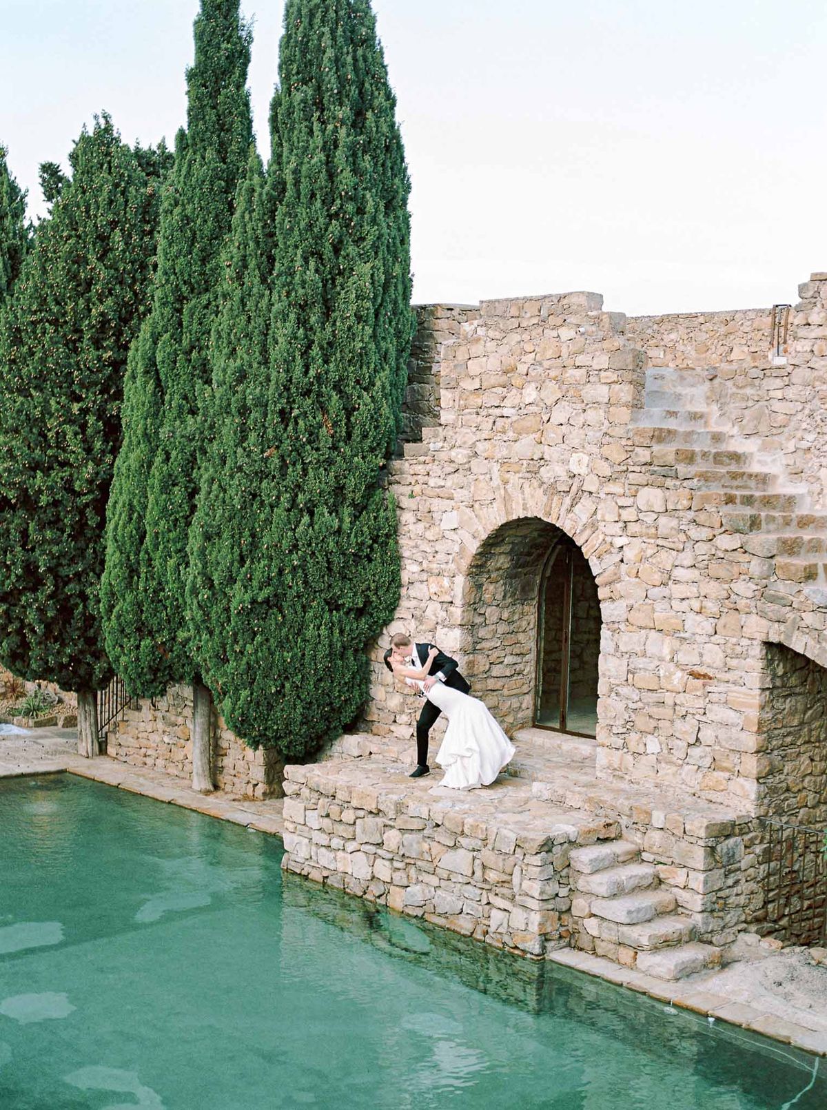 Groom dips bride for kiss beside turquoise pool with limestone arched doorway and cypress trees beyond