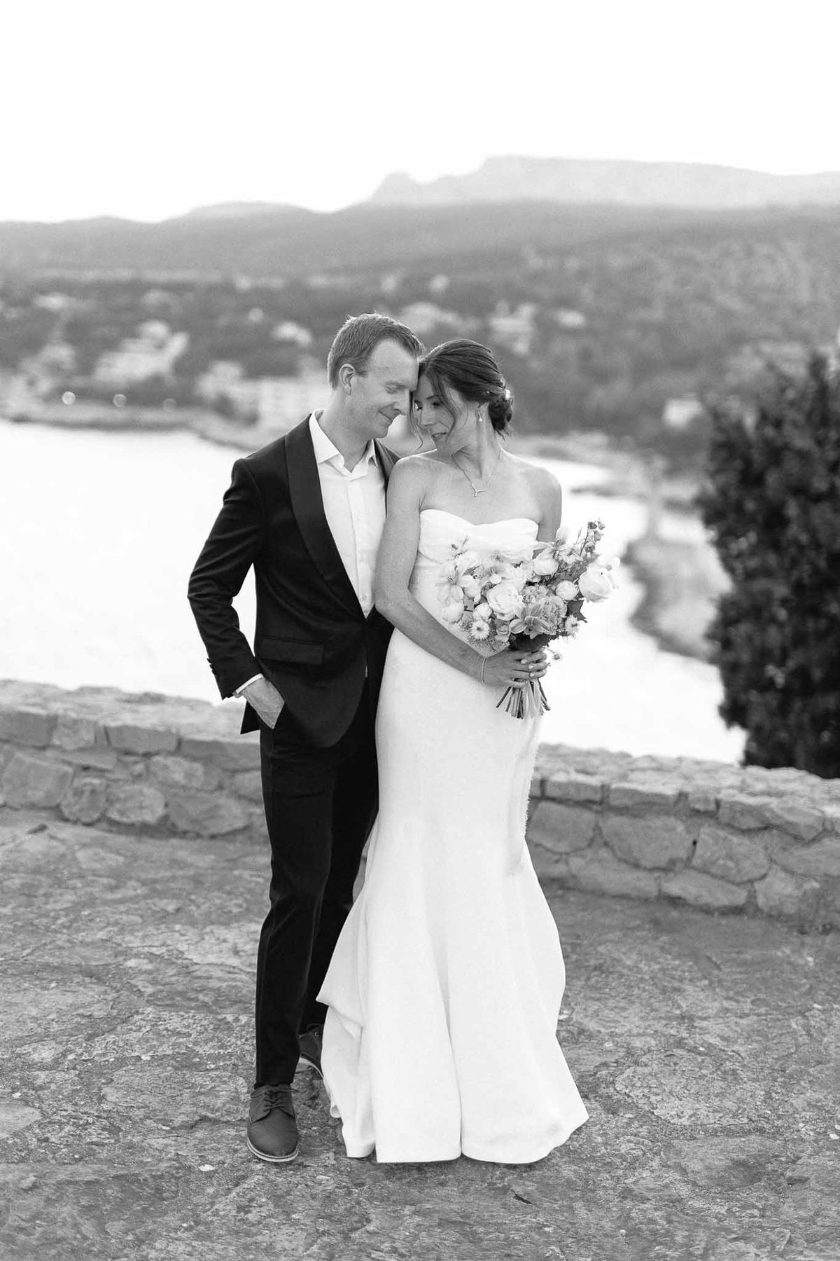 Black-and-white portrait of bride and groom on stone terrace overlooking coastal bay and mountains