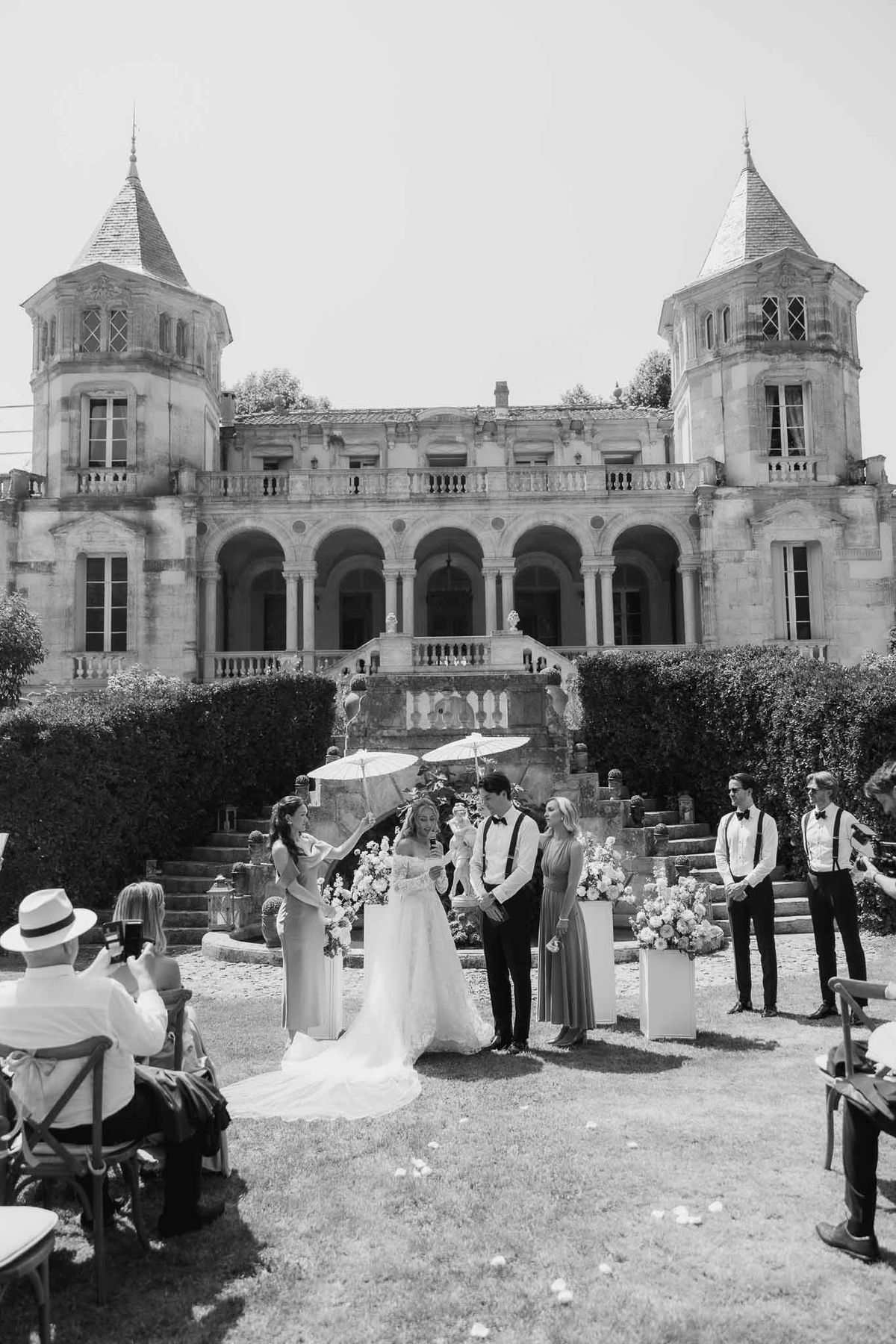 Black and white ceremony at twin-turreted chateau with parasol, pedestals, and groomsmen in suspenders