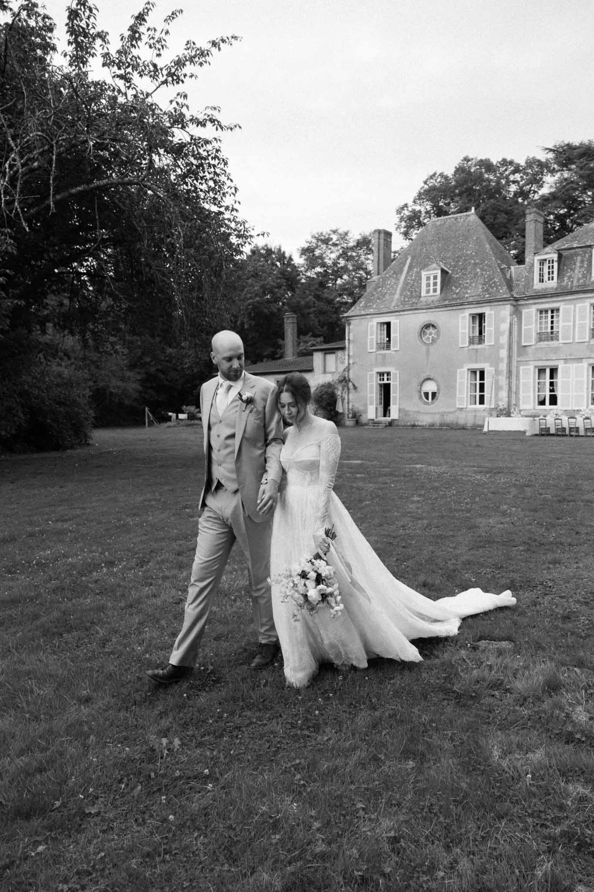 Black and white couple walking across lawn holding hands with flowing lace train and chateau behind