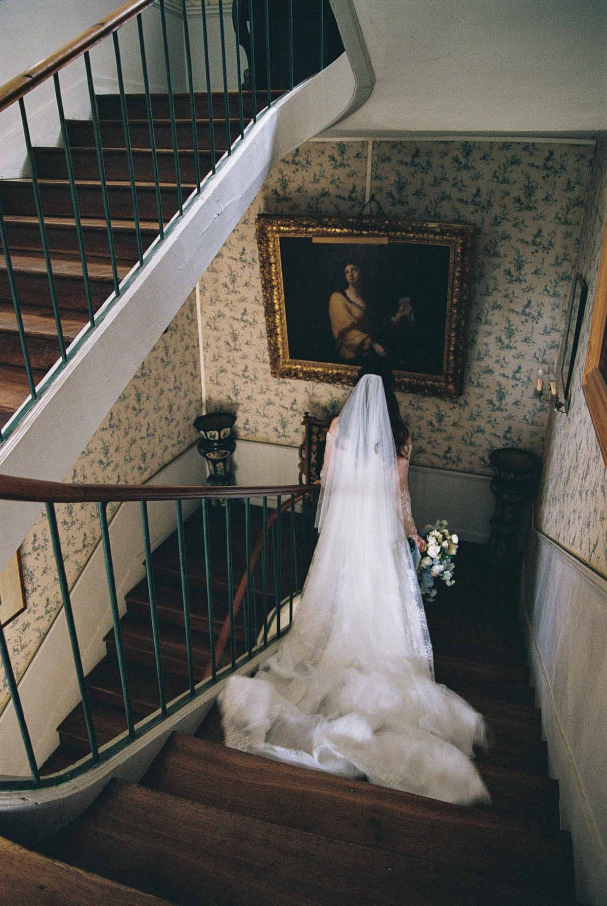 Bride with cathedral-length veil cascading down chateau staircase with toile de Jouy wallpaper and gilt-framed painting