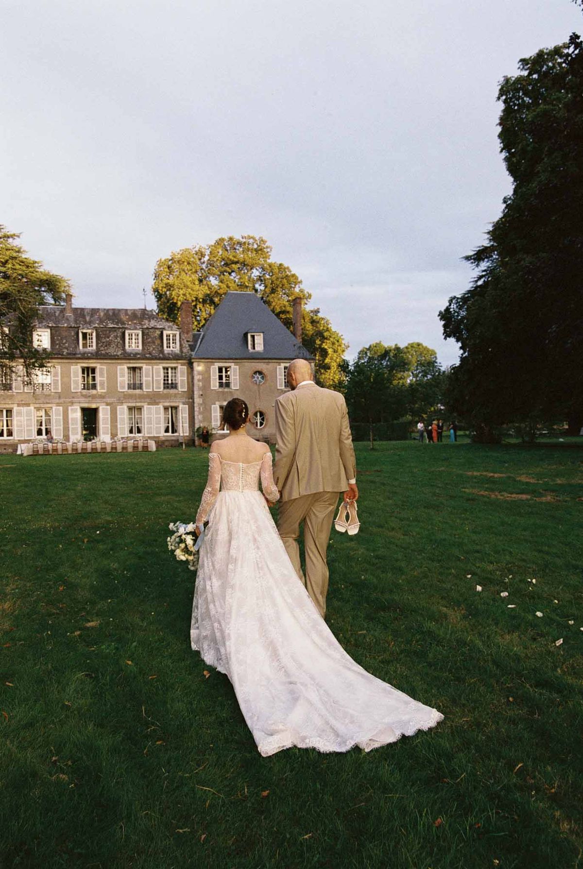 Bride and groom walking hand-in-hand across lawn toward French chateau, groom carrying bride's shoes