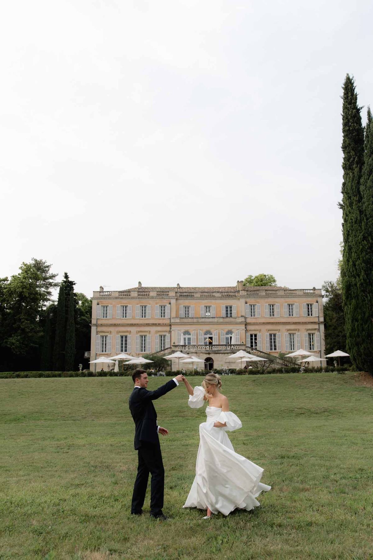 Groom spinning bride in puff-sleeve gown on lawn before ochre chateau with market umbrellas and cypress
