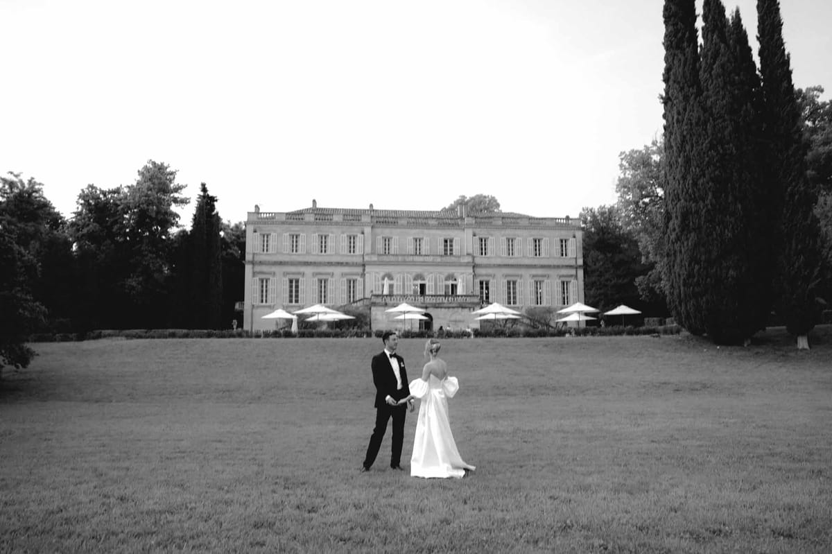 Couple holds hands on lawn before symmetrical French chateau with parasol-lined terrace in B&W
