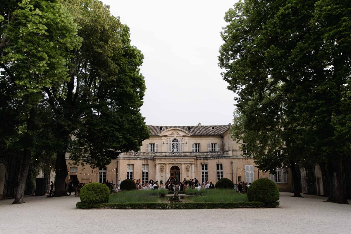Evening reception at round tables before neoclassical chateau with string lights, parterre garden, and fountain