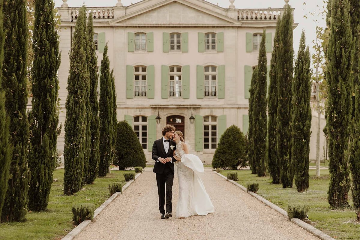 Bride and groom walk cypress-lined allee toward pink stone chateau with sage shutters and pediment
