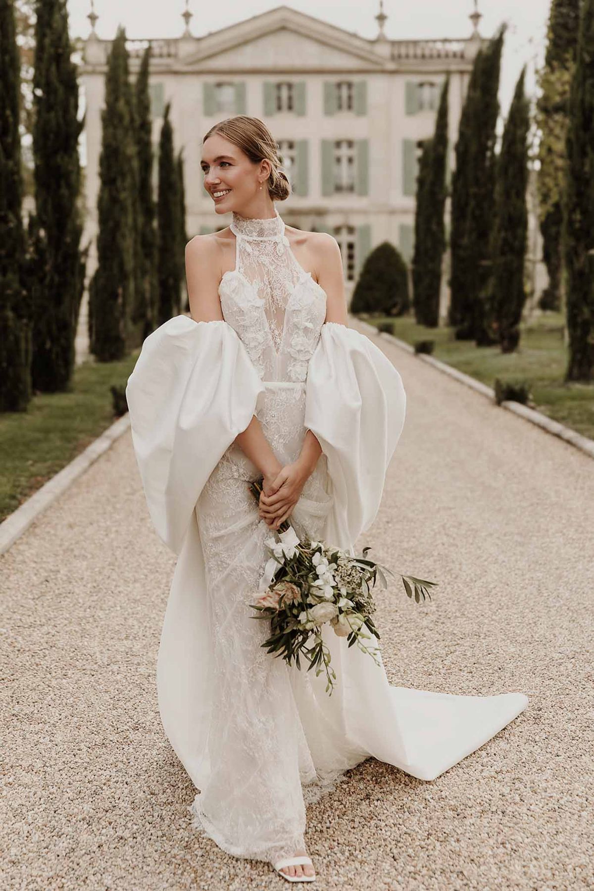 Bride in ivory lace halter gown with white structured cape holding bouquet on chateau gravel driveway
