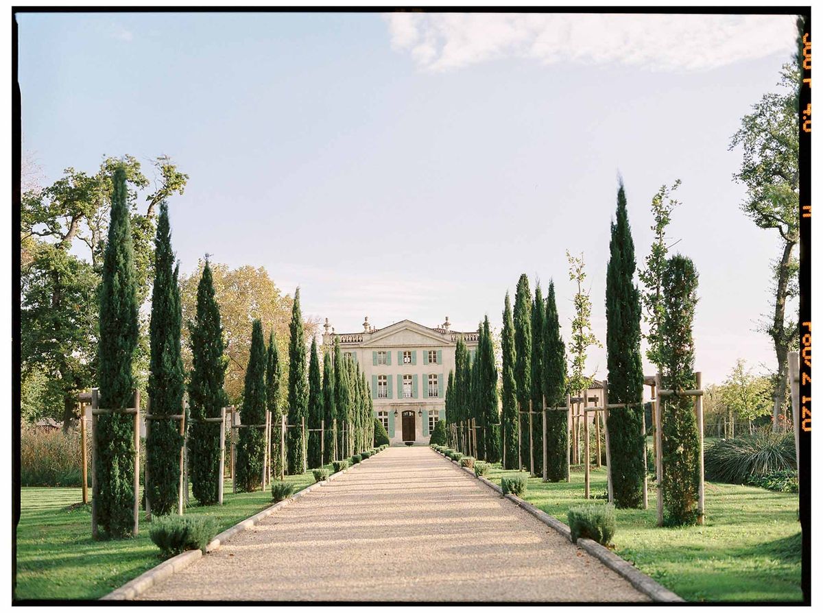 Film shot of cypress-lined gravel driveway leading to classical chateau with pediment and stone urns