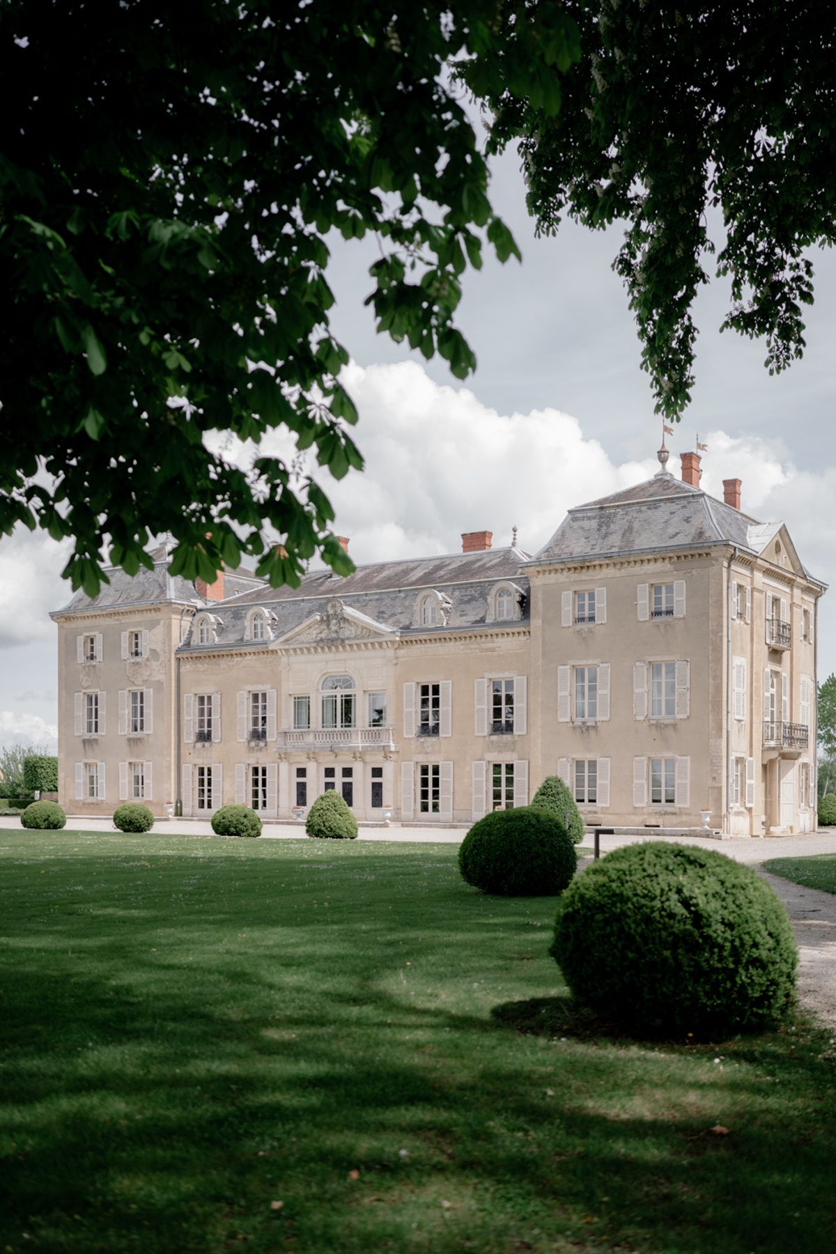 Beige stone chateau with slate mansard roof, red brick chimneys, and round topiary on formal lawn