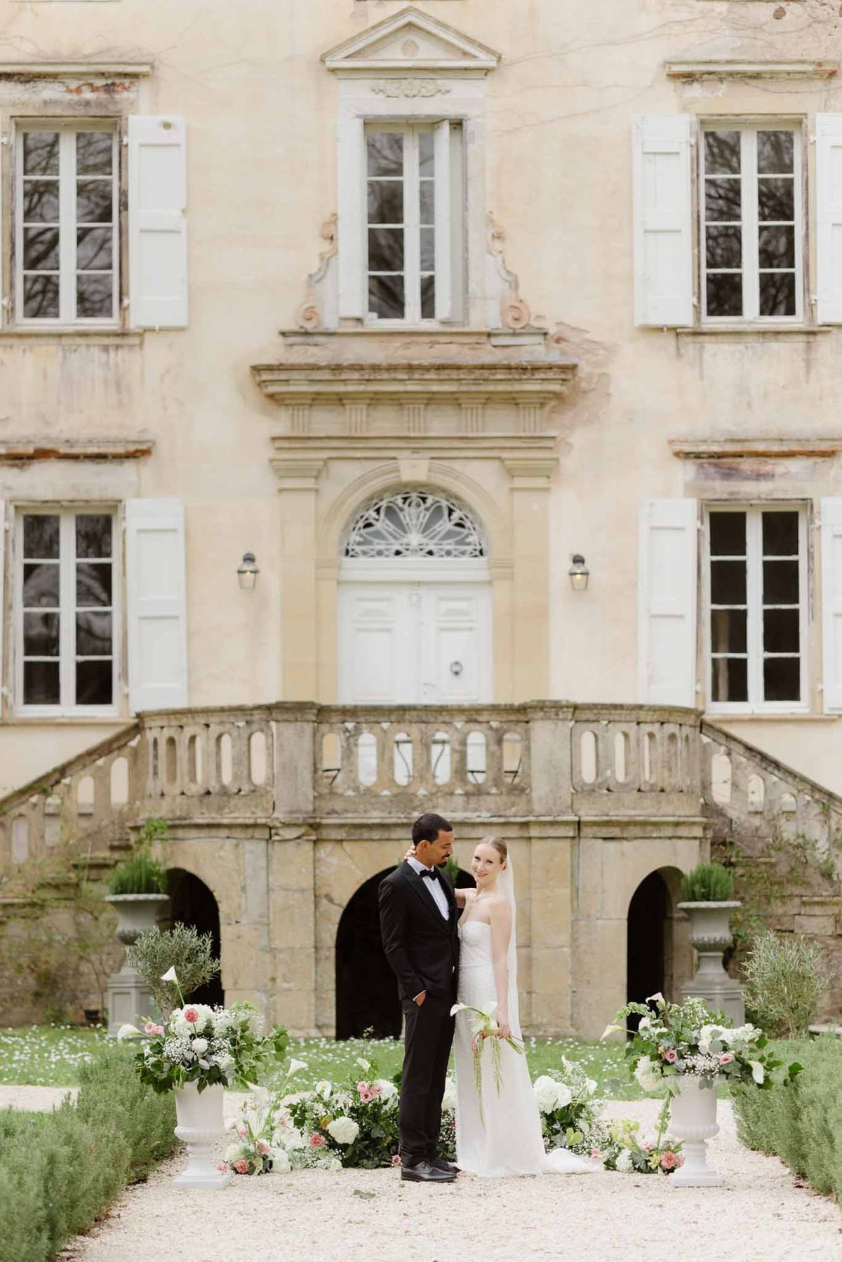 Bride and groom standing before a French chateau facade surrounded by white and blush floral arrangements
