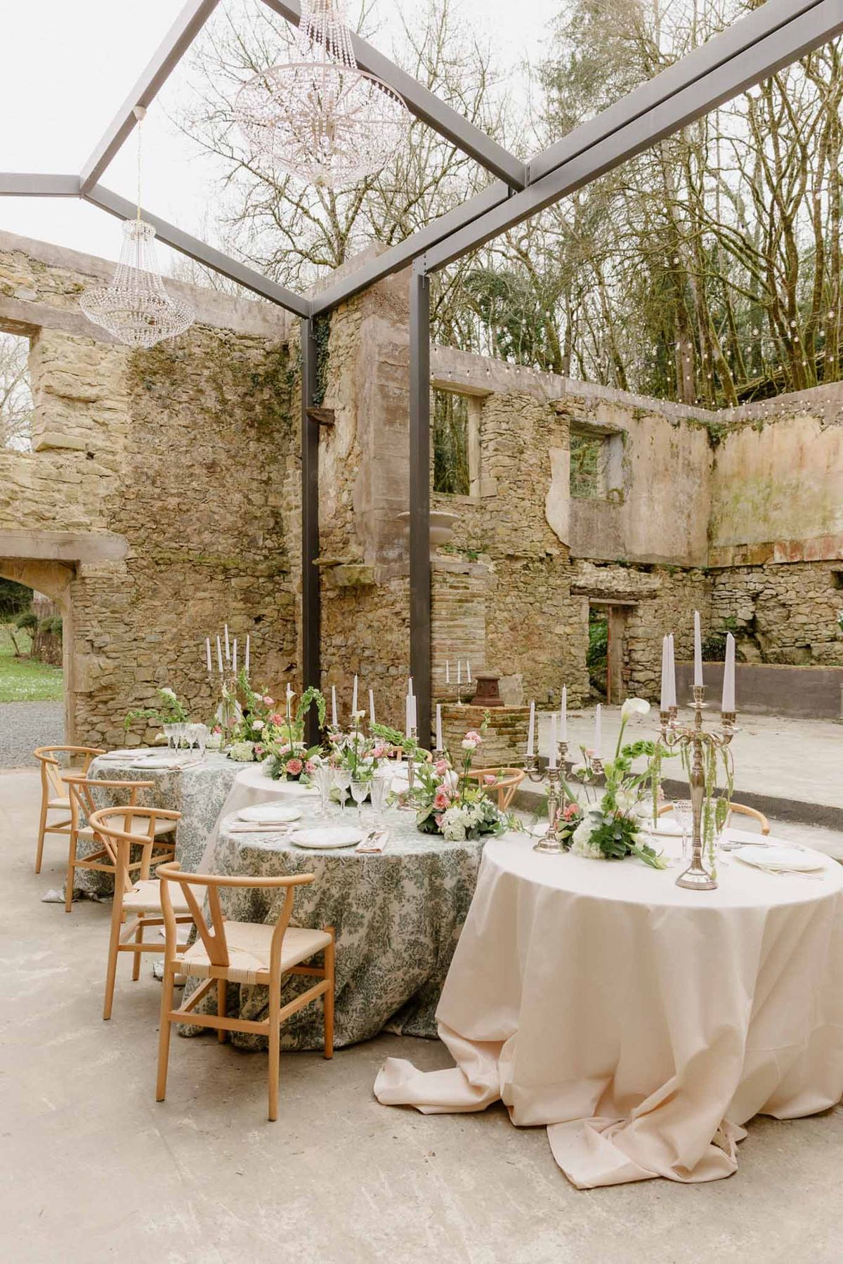 Two round reception tables styled with sage and blush linens under crystal chandeliers in roofless stone ruins