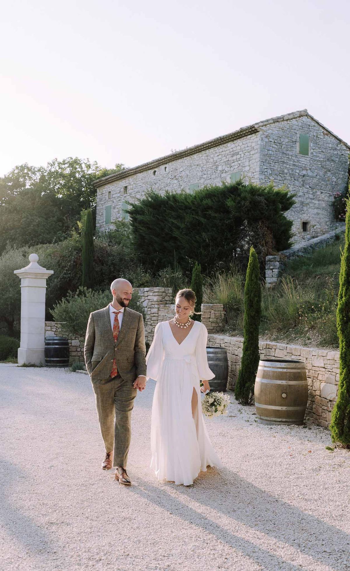Bride and groom walking hand-in-hand along gravel drive past stone farmhouse with green shutters at golden hour