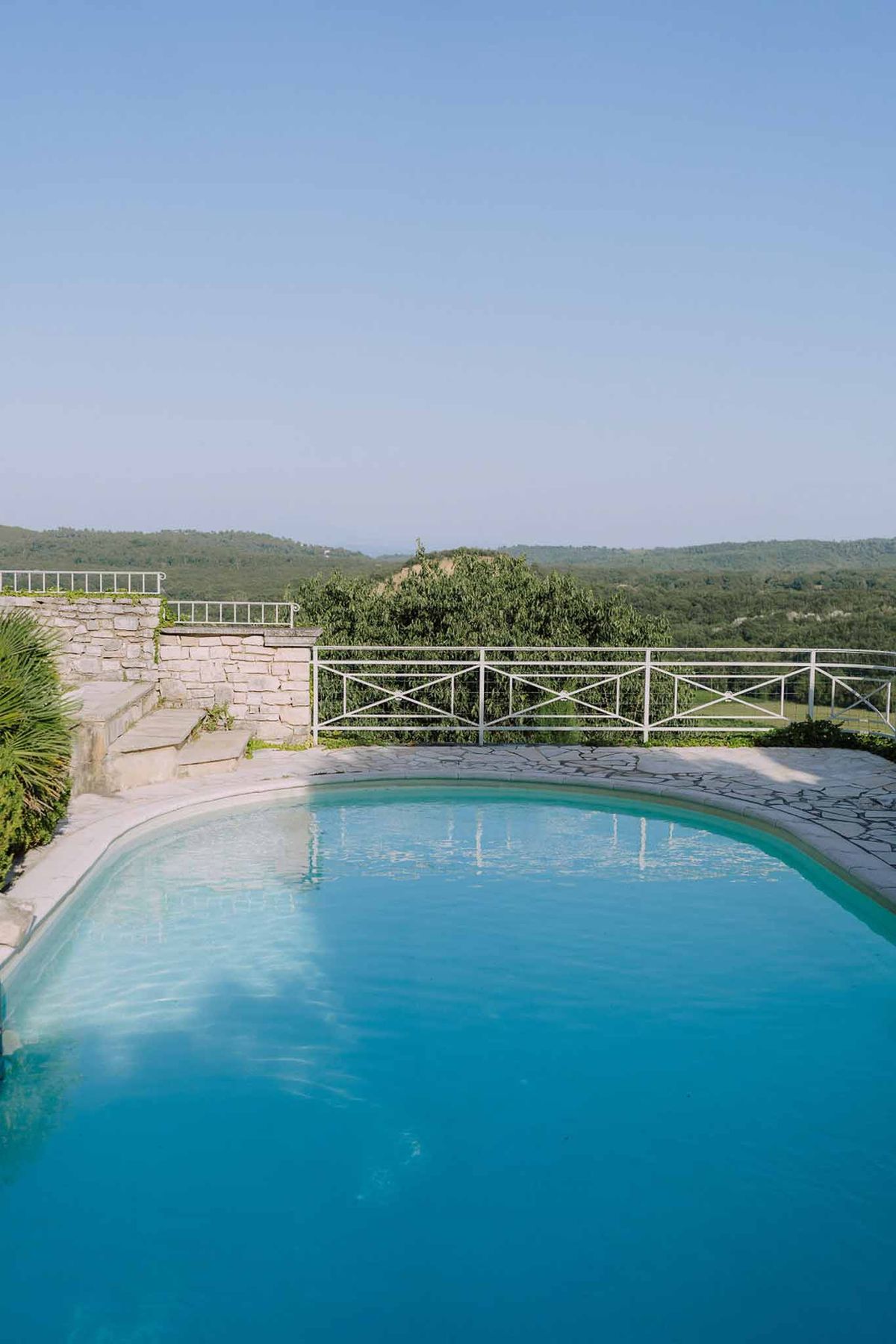 Curved turquoise pool with mosaic tile patio and white metal railing overlooking panoramic hillside countryside
