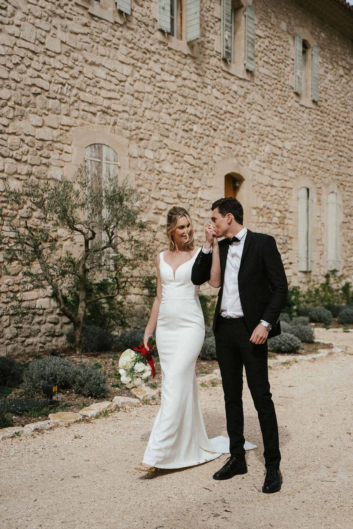Groom kissing bride's hand beside limestone chateau, bride holding white ranunculus bouquet with red ribbon