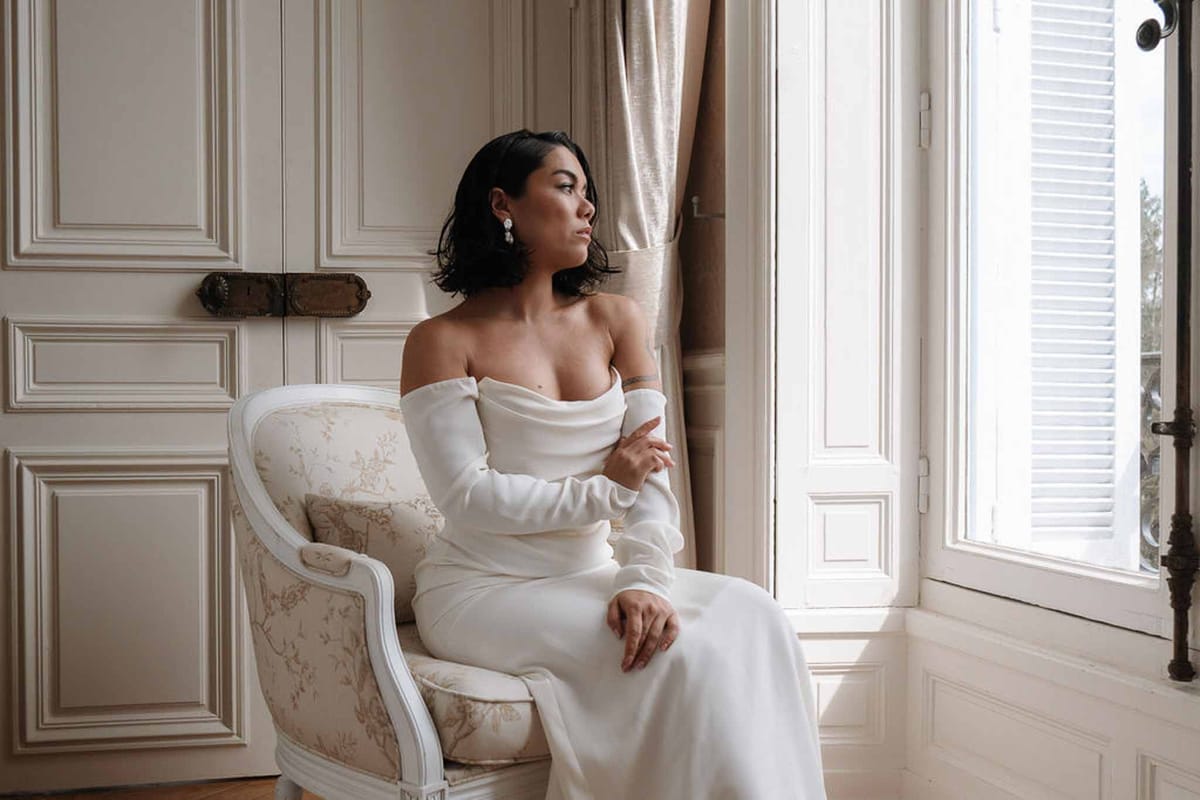 Bride in off-shoulder long-sleeve gown seated in Louis armchair by shuttered window