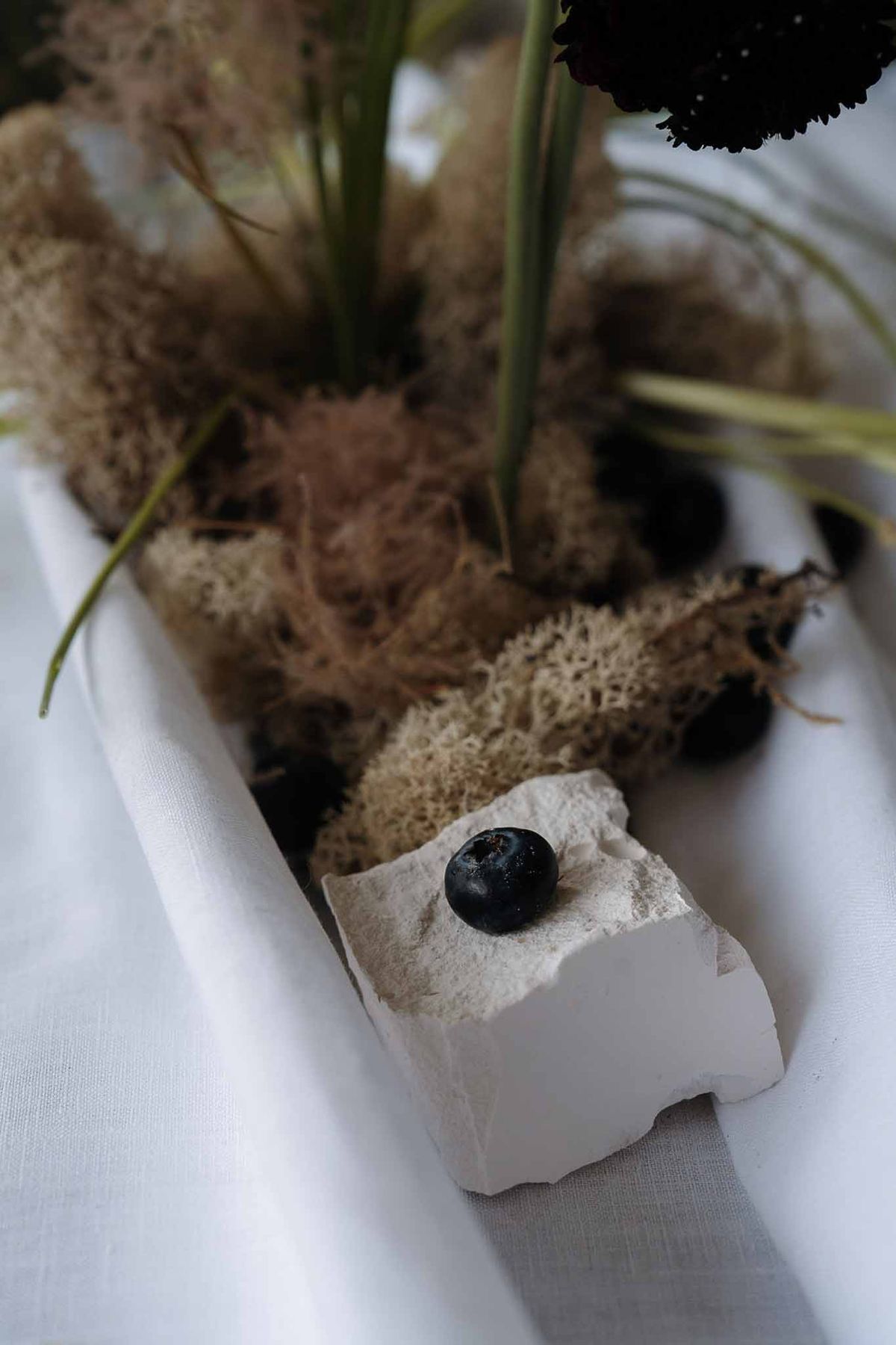 Moody table centerpiece with dried moss, dark stones, green stems, and burgundy scabiosa flower