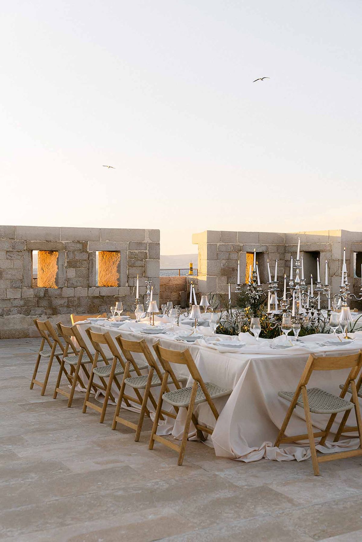 Outdoor reception table on stone terrace with silver candelabras and coastal view at sunset
