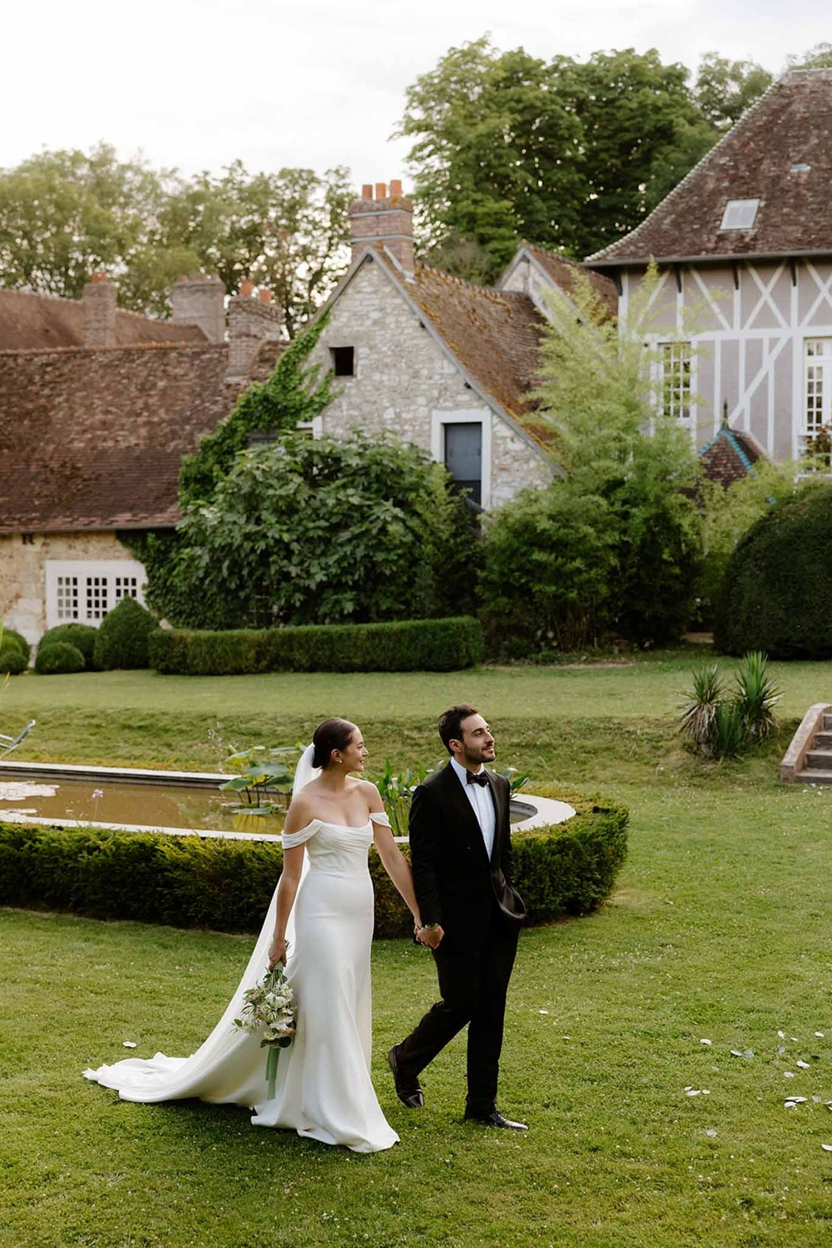 Couple walking on lawn with white ranunculus bouquet before Norman estate with lily pond and half-timbered facade