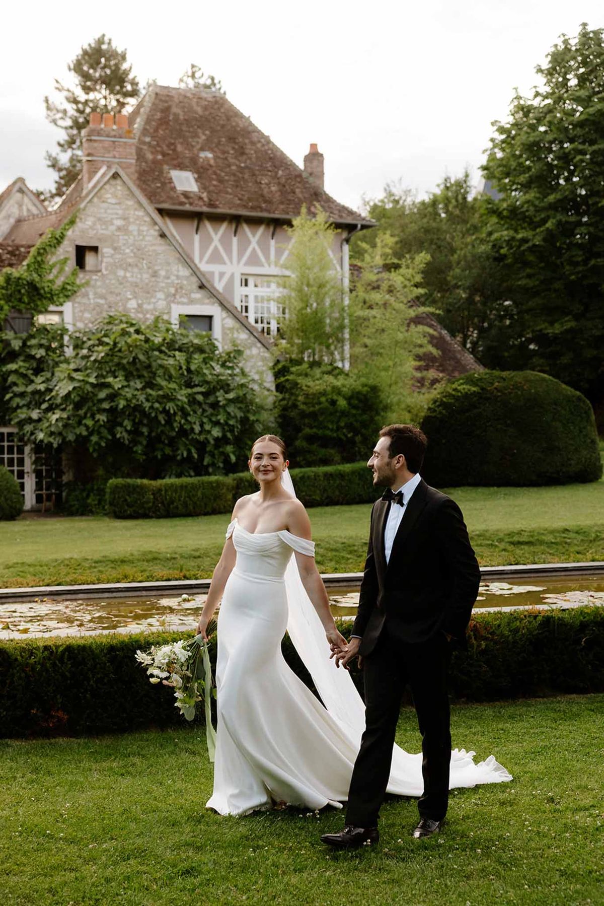 Couple walking on lawn before half-timbered manor with ornamental pool, bride in cathedral veil