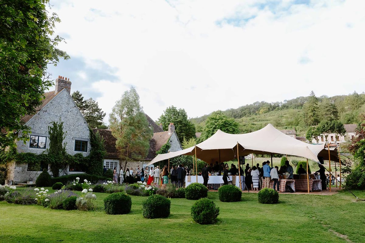Guests mingling under cream stretch tent on lawn before stone Norman manor house with clipped hedges