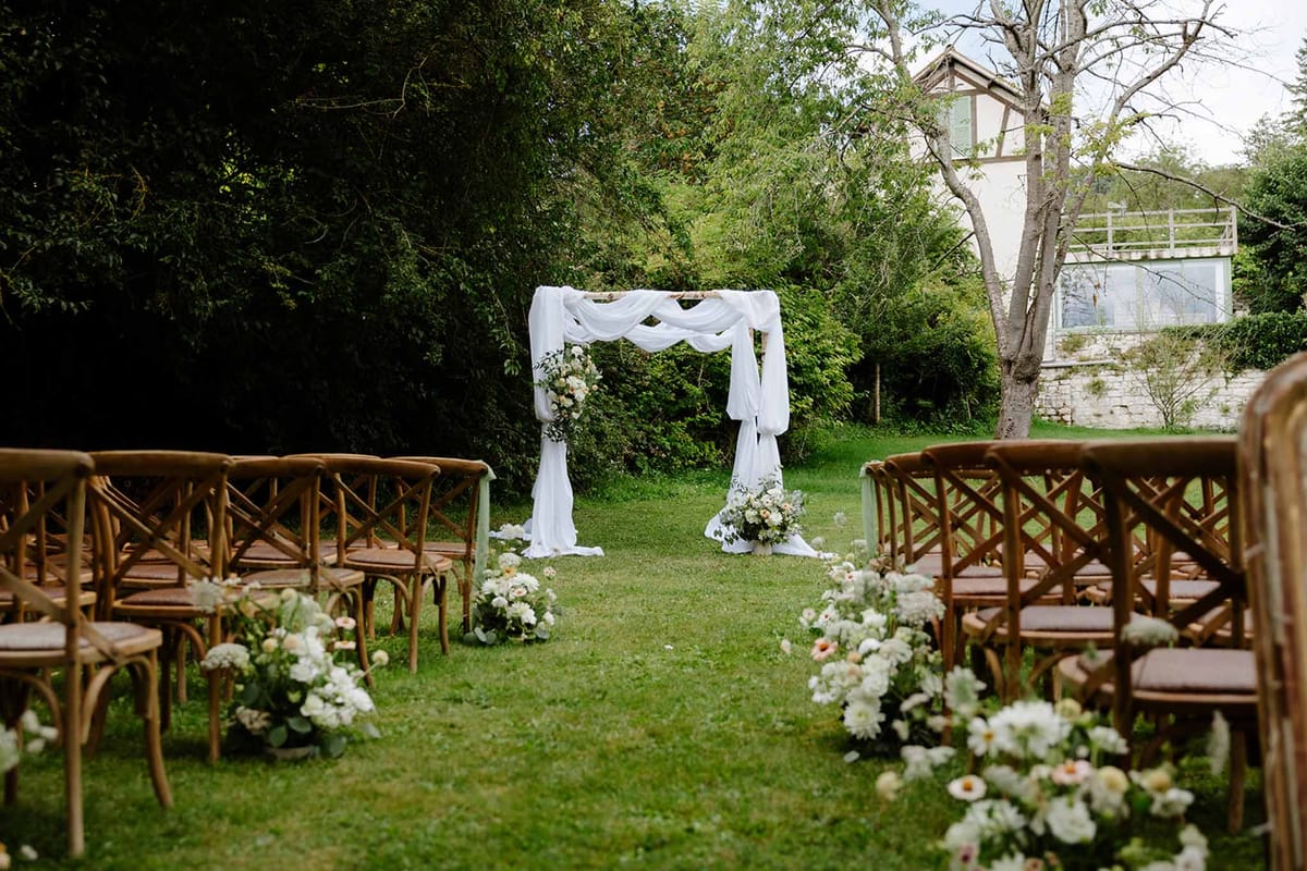 Garden ceremony aisle with fabric-draped wooden arch, cross-back chairs, and white dahlia arrangements on lawn