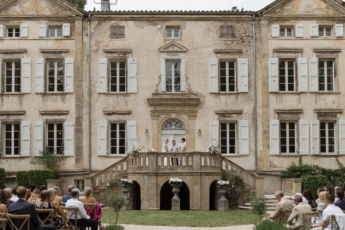 Ceremony on chateau stone staircase with white floral urns and 50 guests seated on courtyard lawn
