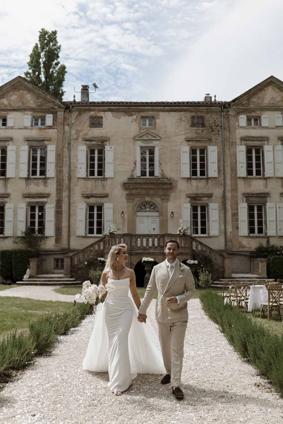 Couple walking on gravel with white peony bouquet and champagne before three-storey stone chateau