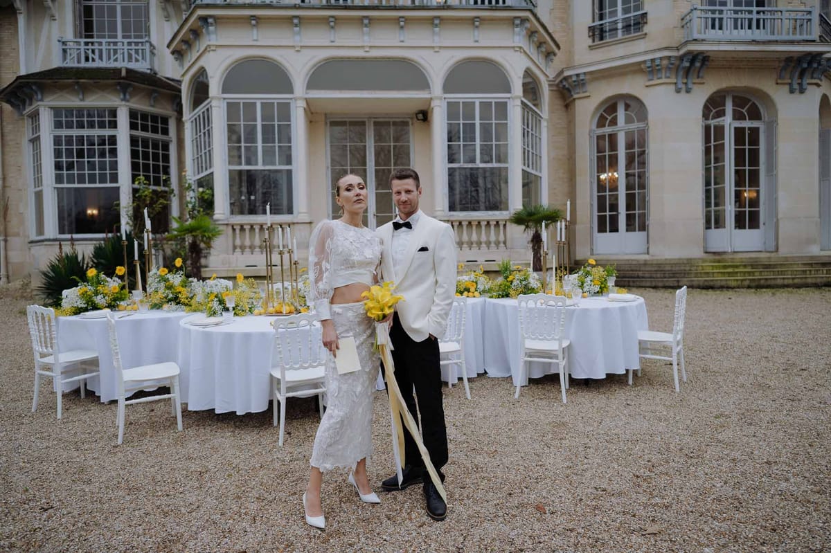 Couple in white attire with yellow calla lily bouquet in front of styled reception tables