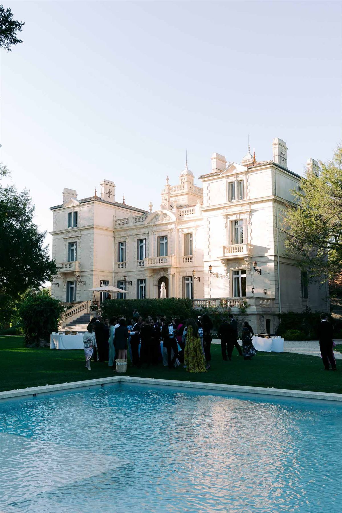 Golden hour cocktail reception on chateau lawn with guests in formal attire gathered beside rectangular pool
