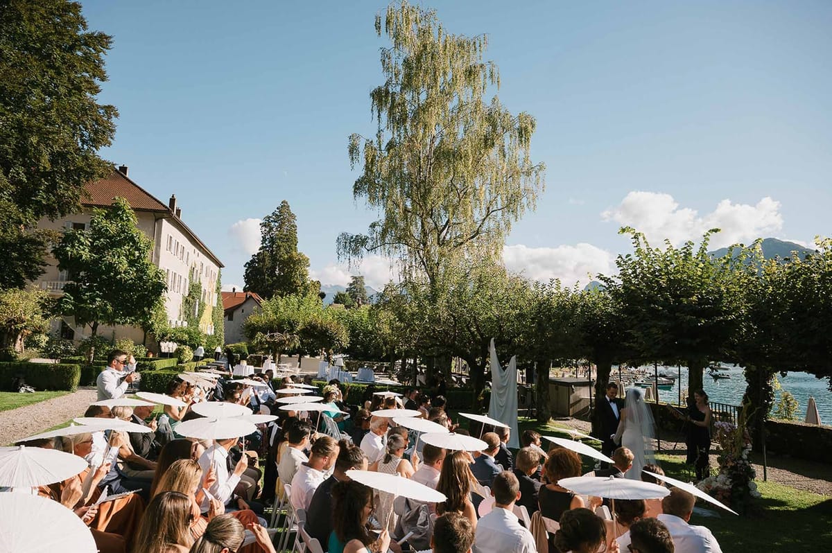 Guests with parasols watching ceremony on lakeside lawn at Abbaye de Talloires