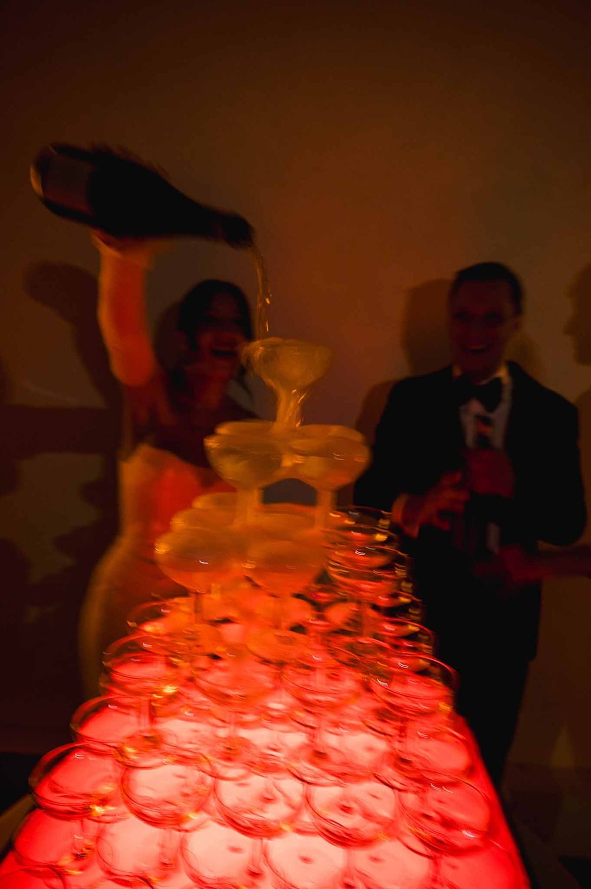 Bride pouring champagne into red-lit glass tower at reception at Abbaye de Talloires