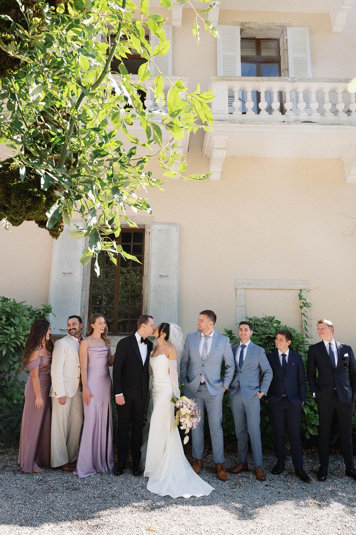 Bride and groom kiss as the wedding party lines up against the abbey facade at Abbaye de Talloires