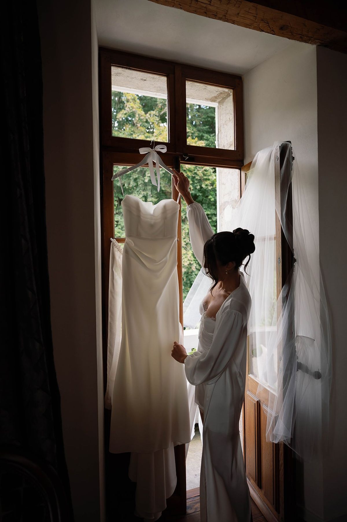 Bride lifts her satin gown beside cathedral veil at the abbey window at Abbaye de Talloires