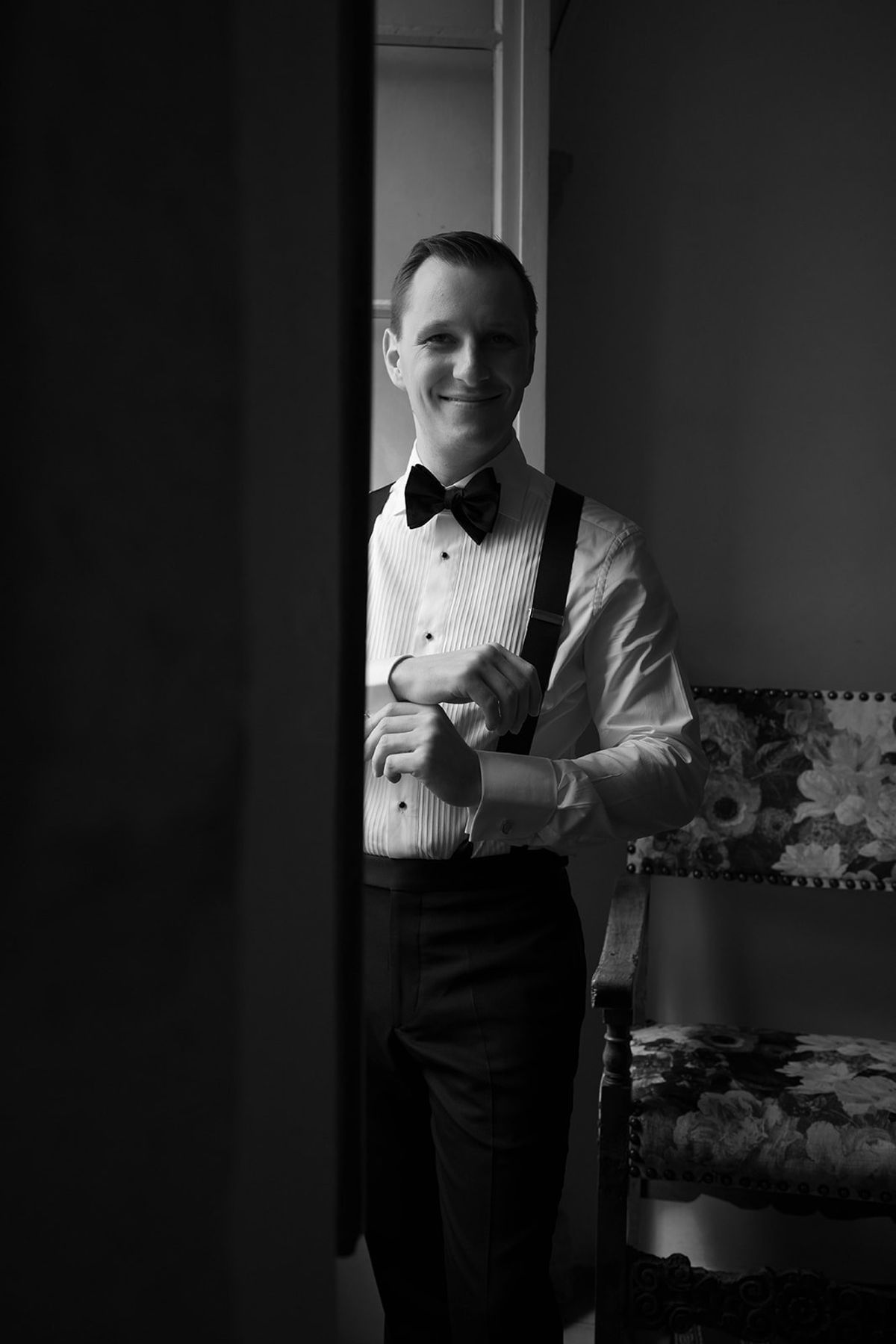 Groom in bow tie and suspenders adjusts his cuffs by an abbey window at Abbaye de Talloires