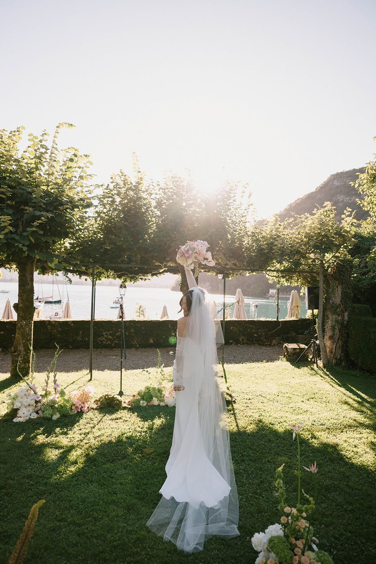 Bride raises her bouquet toward Lake Annecy at golden hour at Abbaye de Talloires