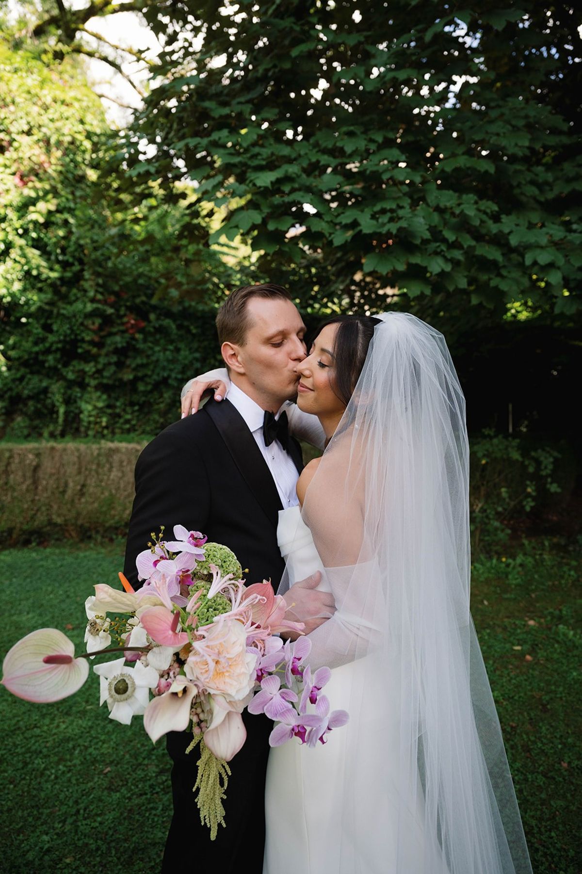 Groom kisses bride's cheek as she holds her cascading orchid bouquet at Abbaye de Talloires