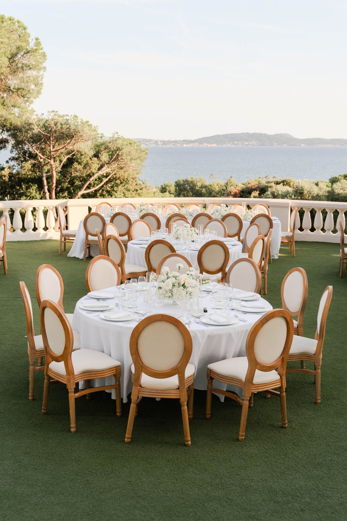 Round dining tables with white linens and rose centerpieces on seaside lawn terrace with balustraded railing