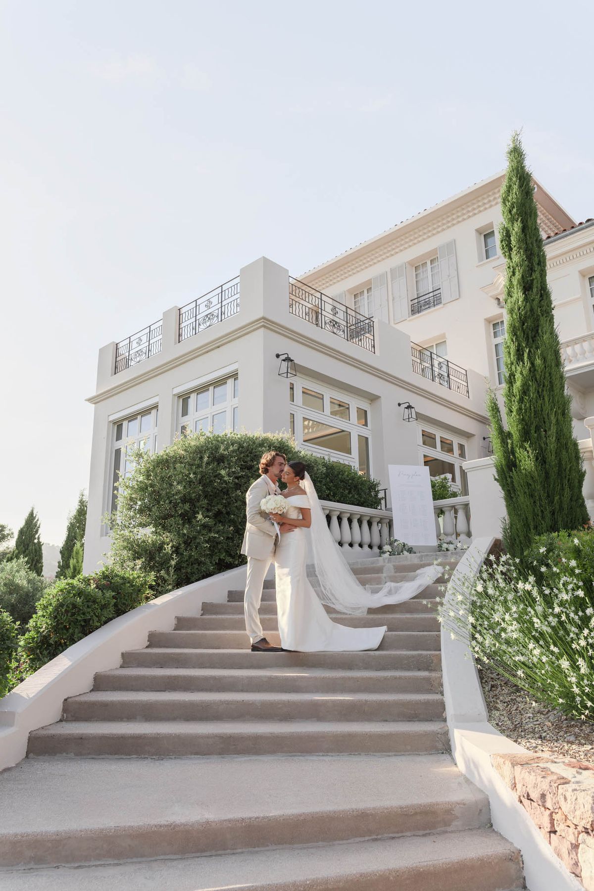 Bride and groom embracing on stone staircase with cathedral veil trailing before white villa
