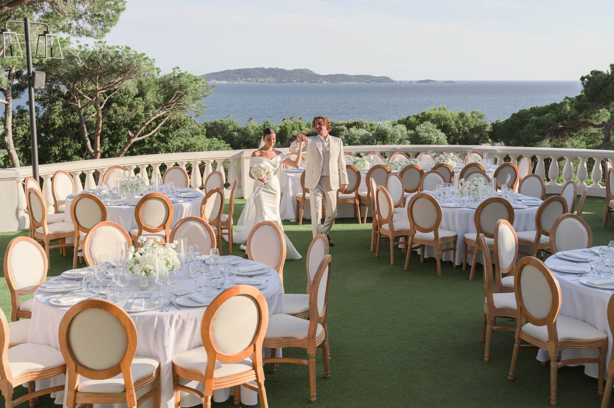 Bride and groom walk through seaside terrace reception with round tables and white floral centerpieces