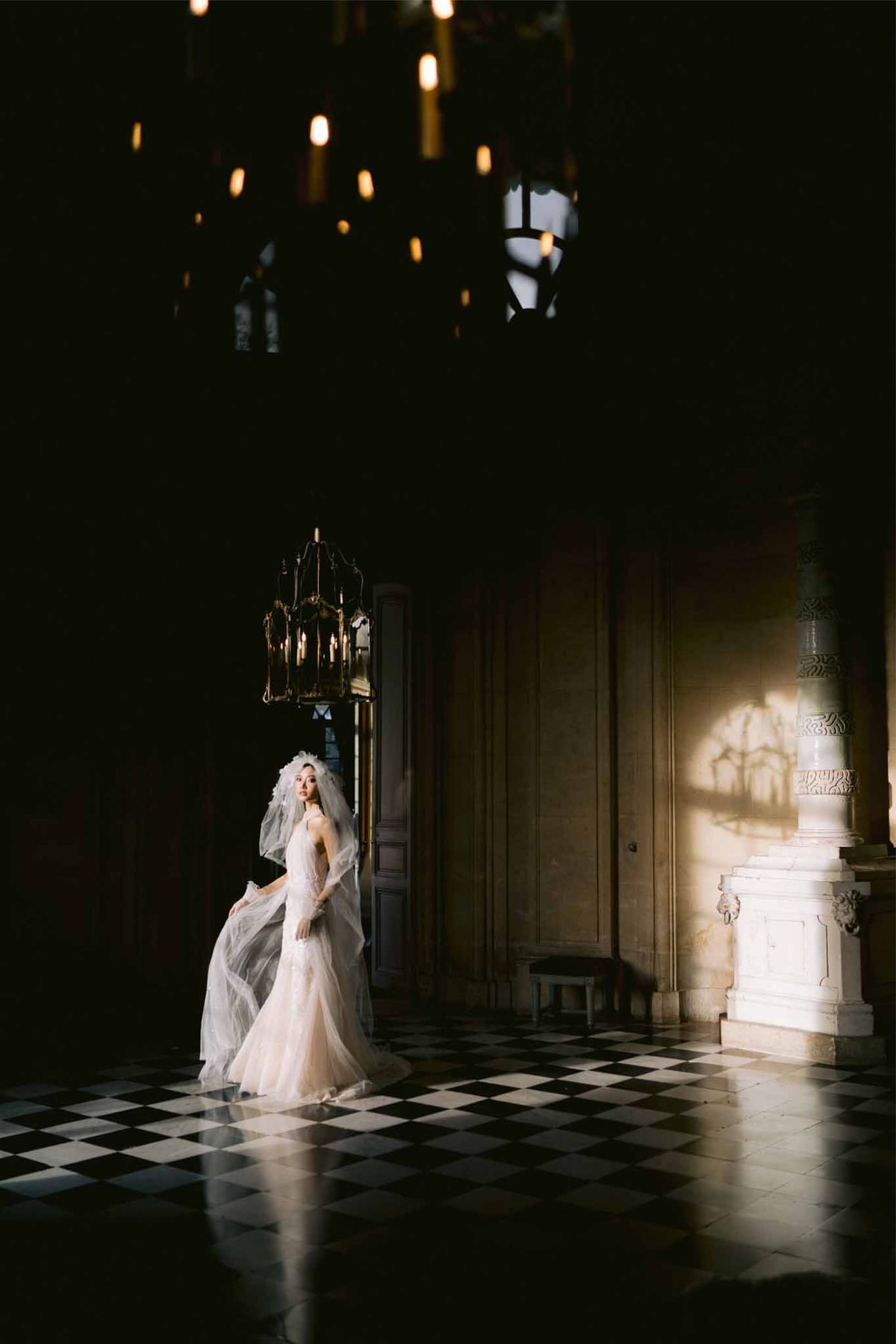 Bride with cathedral veil on diamond marble floor in dramatic chateau hall with brass chandeliers and shadow
