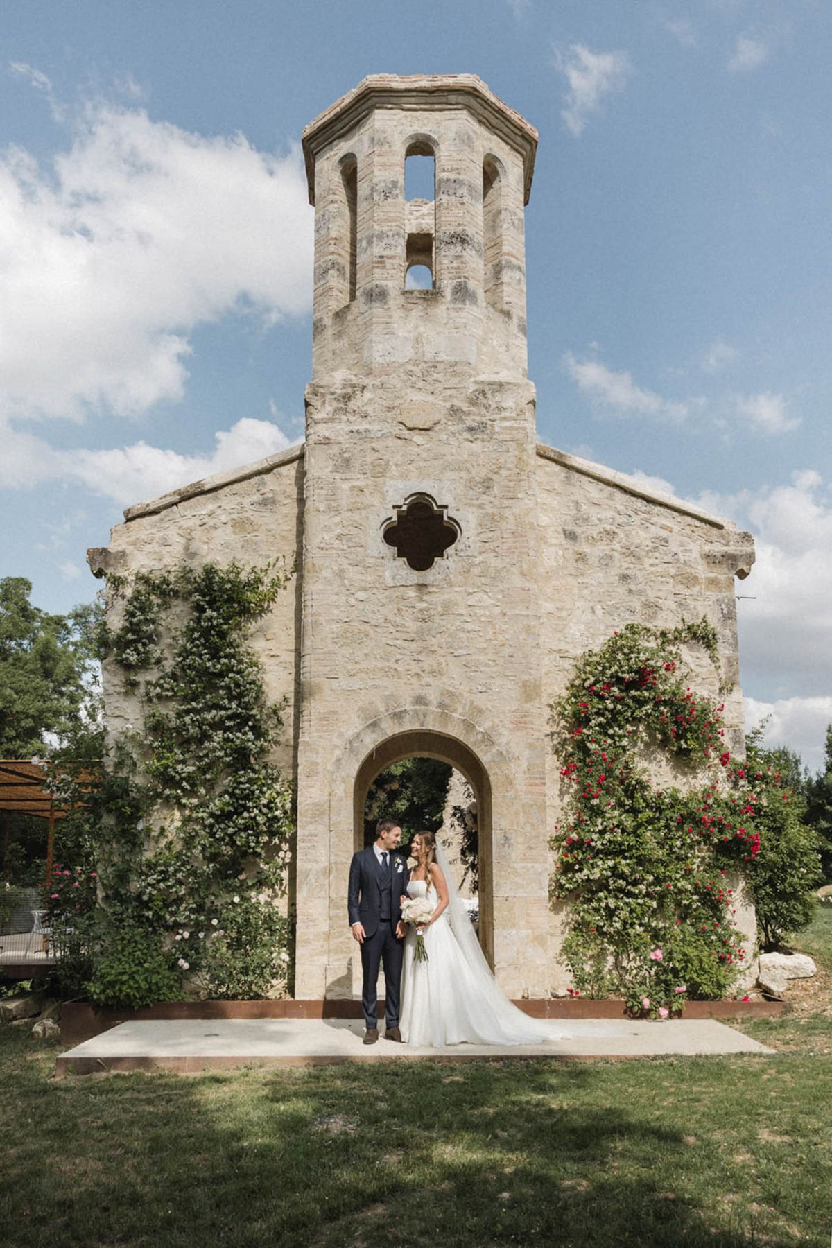 Bride and groom smiling at each other in front of a Romanesque stone chapel with climbing roses