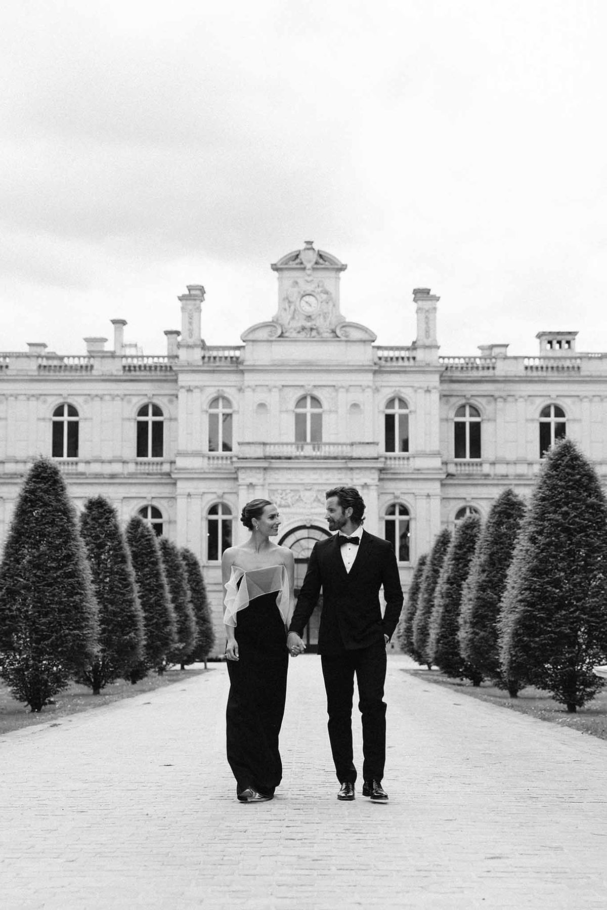 Black and white couple on cobblestone allee between conical topiaries before ornate chateau facade