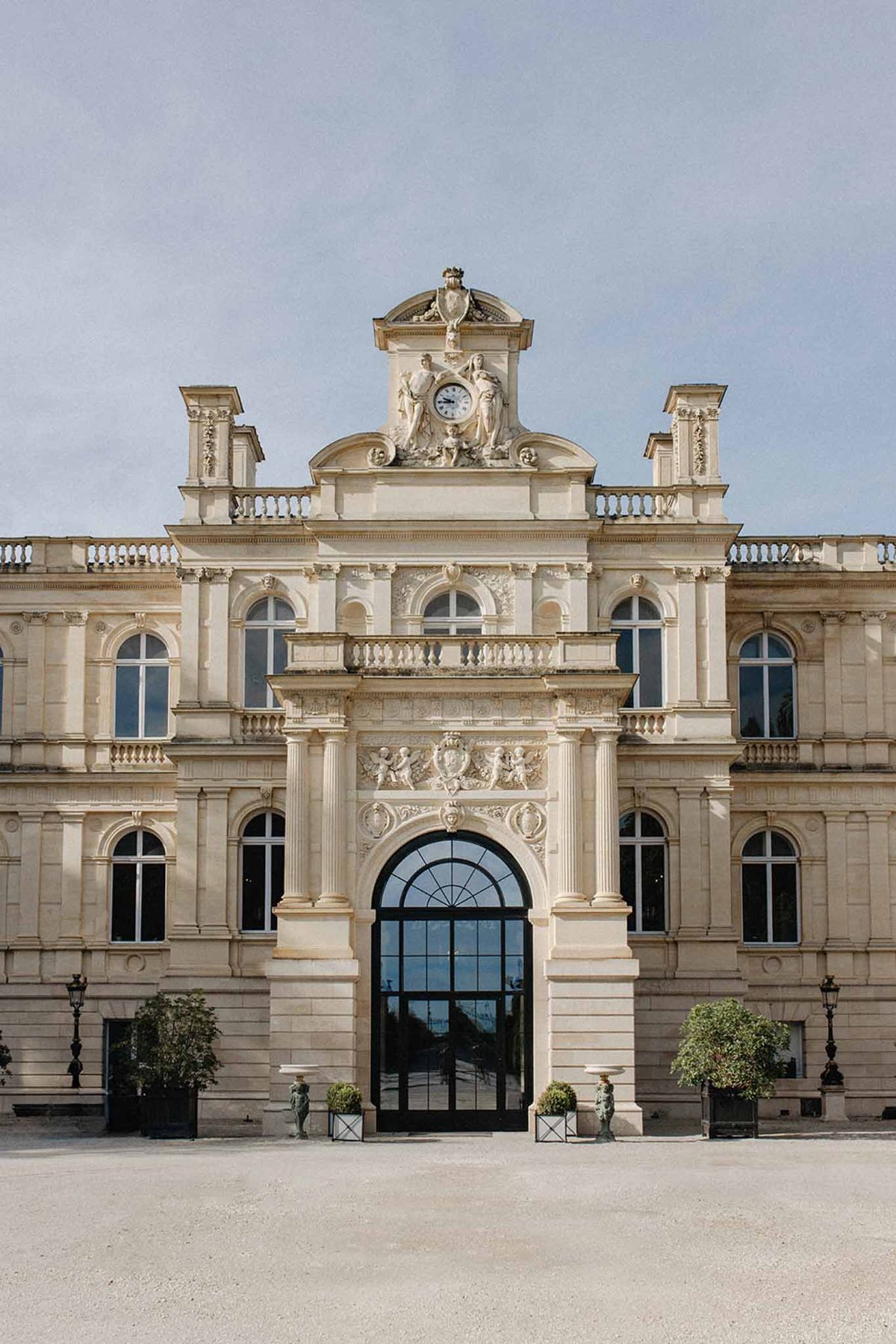Classical cream stone chateau facade with clock pediment, sculpted figures, arched glass entrance, and topiary planters