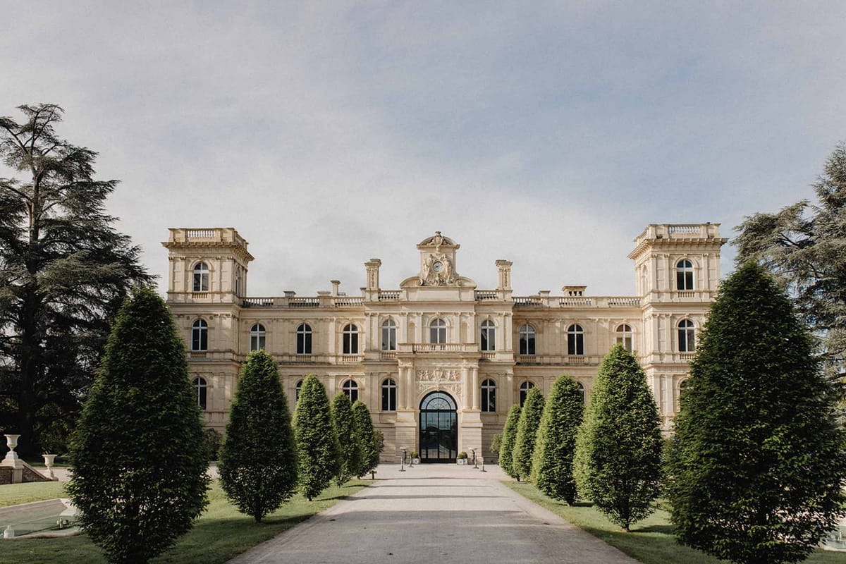 Symmetrical cream chateau with clock cartouche, arched gates, and conical topiary allee on gravel drive