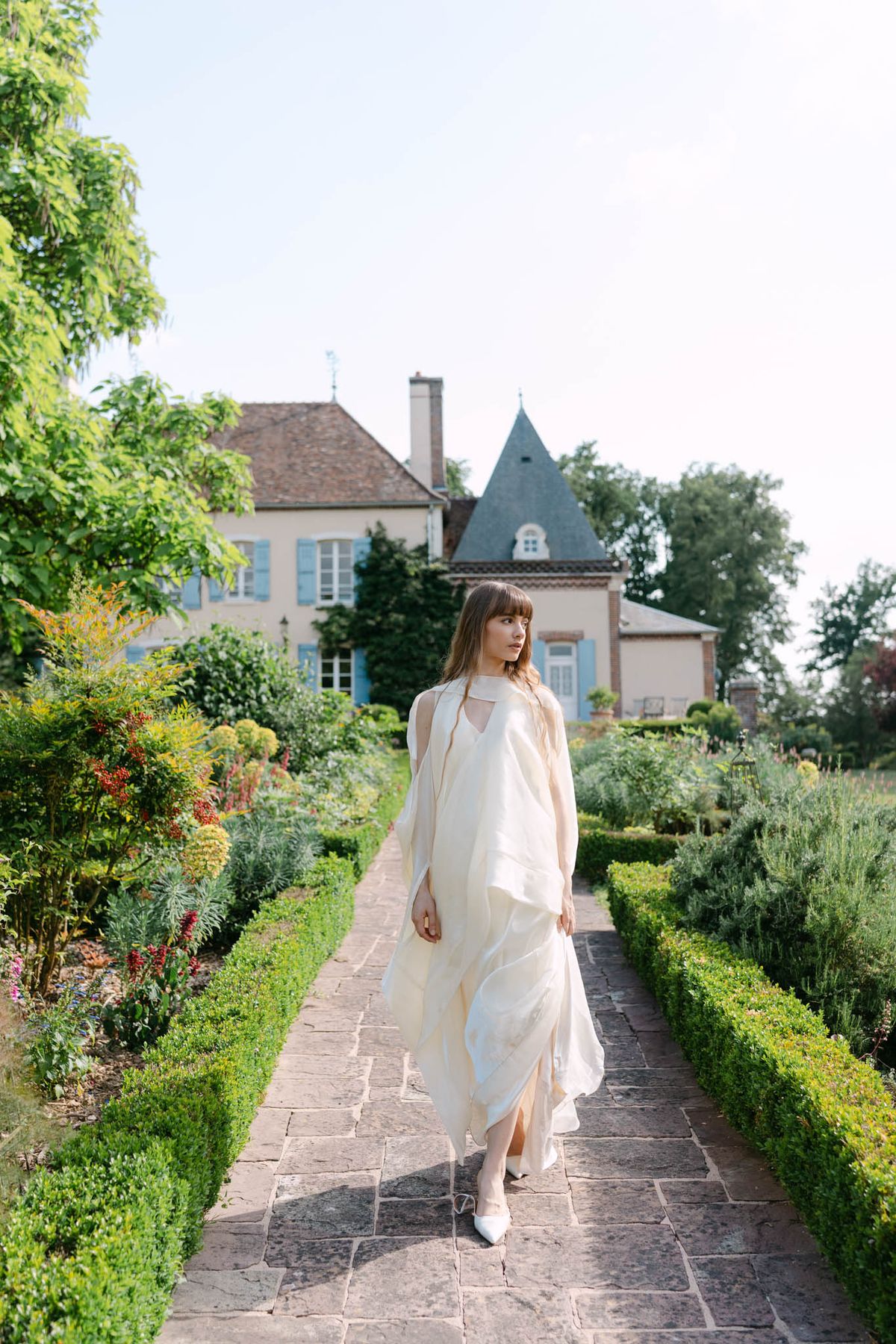 Bride in ivory silk halter gown with sheer wrap on boxwood-lined path before French manor with slate turret
